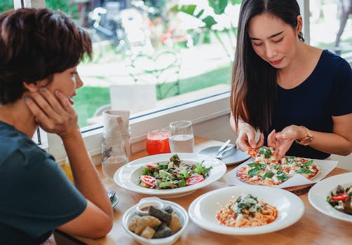 Content Asian woman taking slice of pizza while sitting at table near friend with delicious salad and pasta in restaurant