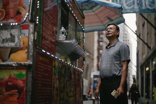 Low angle of pensive male in glasses and casual clothes standing near truck with street food and choosing meal