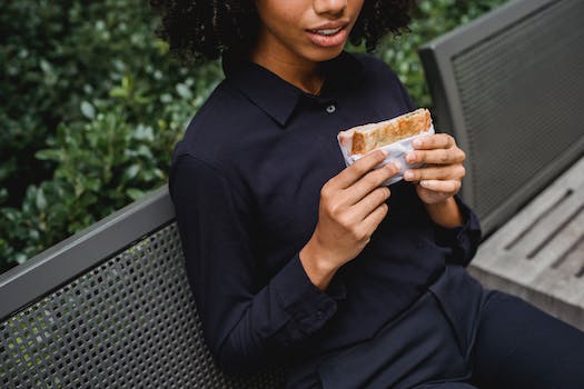 Crop faceless African American female in stylish clothes sitting on bench in park while holding sandwich