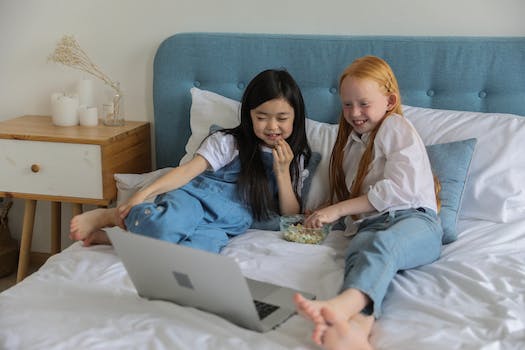 Full body of happy barefooted diverse children with long hairs lying on comfortable bed and eating popcorn while watching funny cartoon on laptop in cozy room