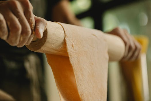 Low angle of unrecognizable person stretching soft dough on rolling pin while preparing food in kitchen