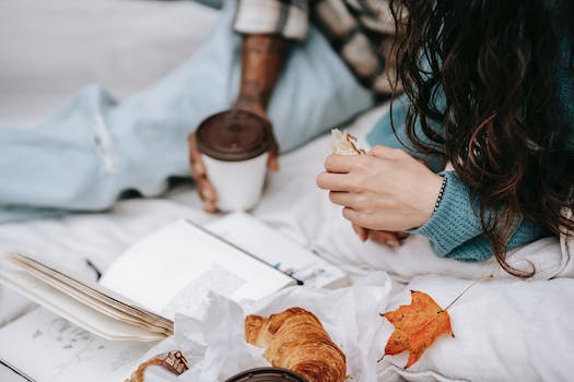Unrecognizable multiracial couple with takeaway hot drink and croissant lying on white plaid with opened book during picnic in autumn day