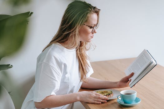 Side view of young blond lady in white t shirt sitting at table with cup of coffee and reading book while spending time at home