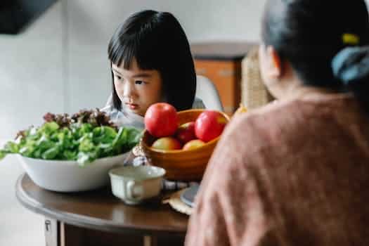 Adorable offended little Asian child eating apples and green healthy lettuce while having lunch with anonymous mother in cozy kitchen