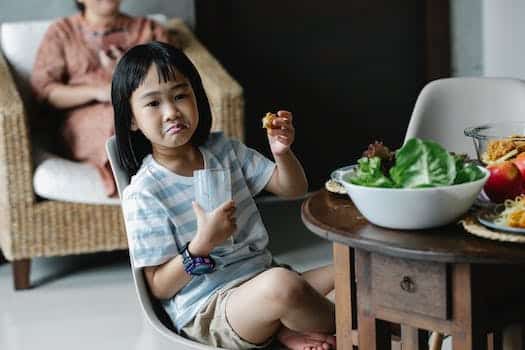 Asian preteen with grimace sitting with glass of milk near wooden table with served food in kitchen during breakfast with blurred background