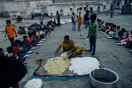 From above of old Indian woman serving food on plates while sitting on ground surrounded with kids in poor area