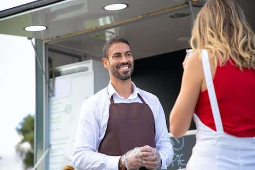 Happy young ethnic waiter looking at anonymous client having lunch near truck cafe