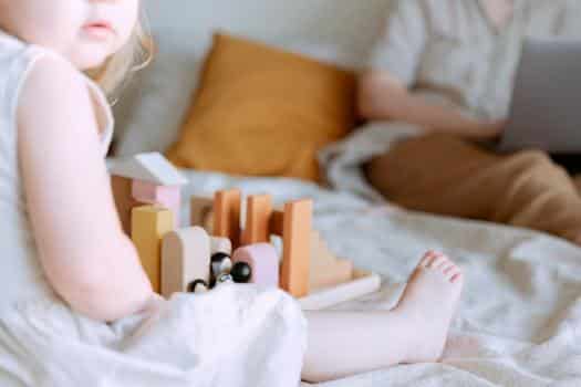 Anonymous toddler girl playing with wooden blocks on bed while mother using laptop