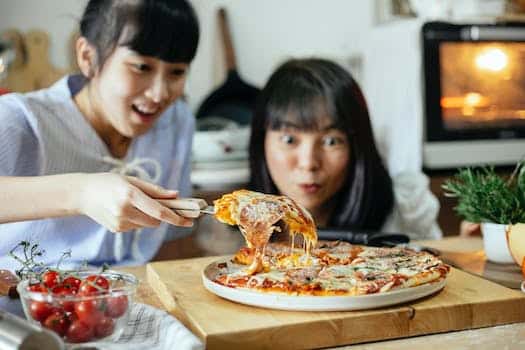 Crop delightful Asian ladies smiling while cutting piece of delicious homemade pizza with stretched cheese on cutting board in kitchen