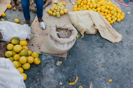 From above of crop faceless seller near pile of ripe oranges on ground and bag of grains at bazaar