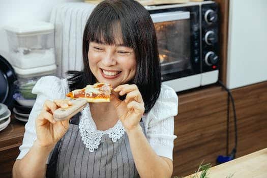 Happy Asian woman enjoying eating slice of cheesy pizza sitting at table in kitchen