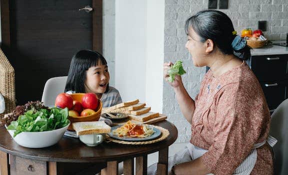 Side view of aged positive Asian woman in apron and dress eating lettuce while having lunch with adorable attentive little granddaughter sitting at table with opened mouth