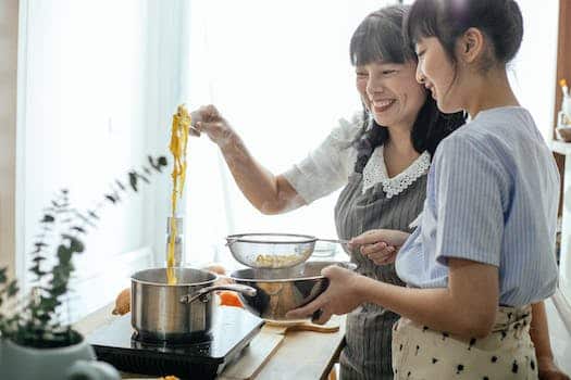 Side view of smiling Asian teenager with middle age mother serving hot boiled pasta in drainer
