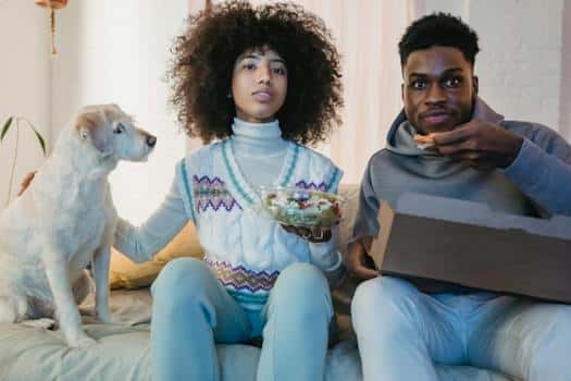 Young ethnic couple watching TV and eating delivered meal sitting on couch near dog