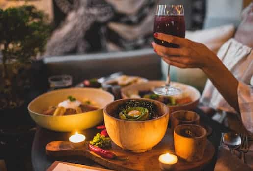 Crop anonymous pregnant female holding glass of red wine while standing near table with lentil dish and salad near hot chili pepper and candles in evening